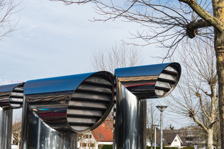 Metal structures are set in a public area. They have curved shapes and gray tones. Trees are visible...