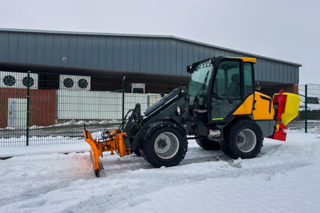 A small snow removal machine is parked on a snowy path near a large storage facility. The vehicle is...