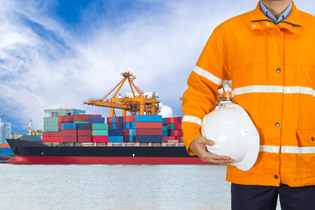 Engineer dockers wearing safety coat and holding a helmet in front of an industrial harbor with cranes and a container ship being unloaded for logistic Import export backgroundの写真素材