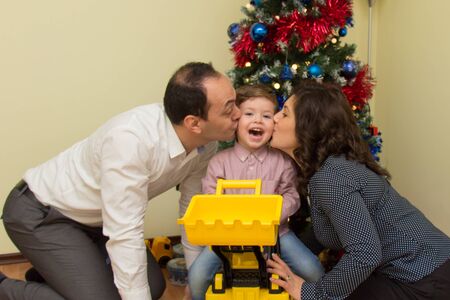 Parents, mother and father, kissing their son on the cheeks with decorated Christmas tree in the backgroundの写真素材