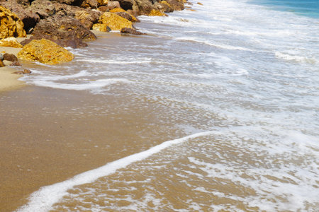 The photo captures a sandy beach as waves crash onto the shore. The foamy waters ebb and flow, leaving behind intricate patterns in the wet sand.の写真素材