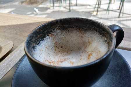 A foamy cappuccino served in a rustic black mug, set against a backdrop of natural light and cafe furniture. the textured froth and earthy tones create a warm, inviting atmosphere ideal for relaxation or reflection.の写真素材