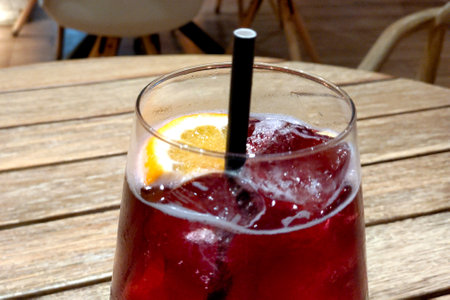 A glass of iced red beverage garnished with a slice of orange and a black straw, sitting on a wooden table in a cafe. the background features blurred wooden chairs, creating a warm, inviting atmosphere.の写真素材