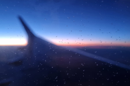 Airplane wing silhouetted against a vivid sunset sky, seen through a window speckled with raindrops. the gradient of colors from deep blues to warm oranges creates a serene atmosphere above the clouds.の写真素材