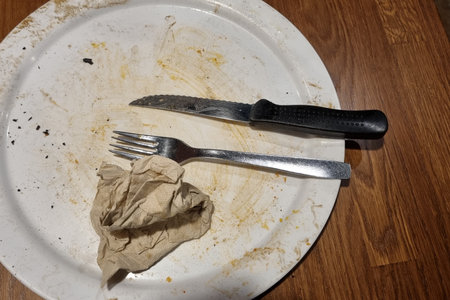 A white plate with food remnants, a crumpled paper napkin, a knife, and a fork rests on a wooden table surface. the image conveys a post-meal scene, emphasizing themes of dining, cleanliness, and domesticity.の写真素材