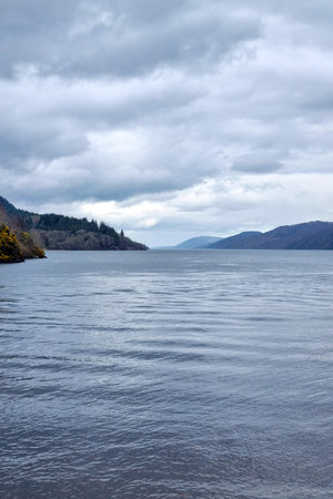 Tranquil lake extending to a mountainous horizon under an overcast sky. the scene is dominated by calm waters, gentle rolling hills, and lush greenery, creating a serene and contemplative atmosphere ideal for nature lovers.の写真素材