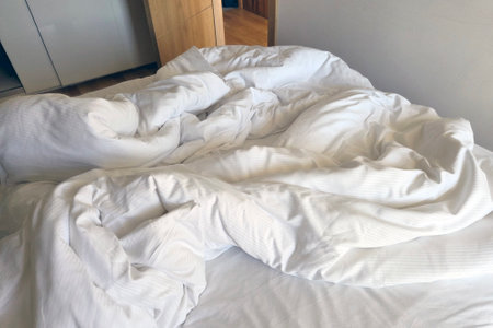 A cozy, inviting unmade bed with crumpled white linens in a sunlit room near a wooden wardrobe. it...