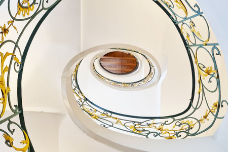 An intricate spiral staircase featuring elegant iron railings adorned with gold floral accents. the upward view emphasizes the curves and symmetry of the staircase, creating a captivating pattern against the white walls.の写真素材