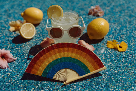 A refreshing summer lemonade garnished with a lemon slice and served in a glass with white sunglasses. set on a sparkling blue surface, surrounded by whole and sliced lemons, vibrant flowers, and a glittery rainbow fan. Generative AIの素材