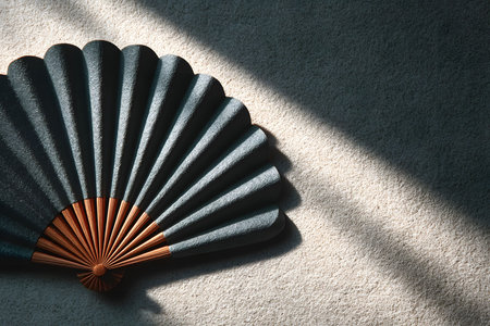 A traditional folding fan displayed against a textured concrete background, illuminated by striking sunlight. the fan's dark fabric contrasts with its wooden ribs, showcasing intricate craftsmanship and cultural elegance. Generative AIの素材