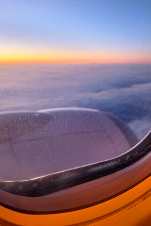 Passengers can see a colorful sunset from an airplane window. The sky shows shades of orange and...