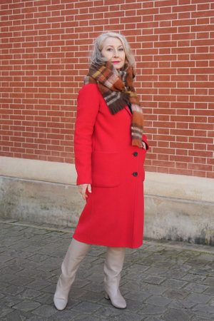 Gray-haired woman of sixty years old in a red coat and a terracotta scarf near a red-orange brick wallの写真素材