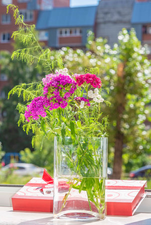 Close-up of a bouquet of carnations on a window on a sunny summer day in a glass transparent vase with a gift box of chocolates.の写真素材