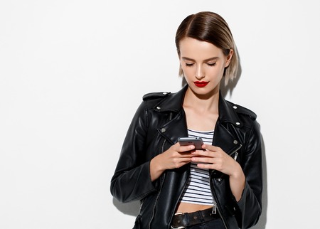 Glamorous young woman in black leather jacket on white background with mobile phoneの写真素材
