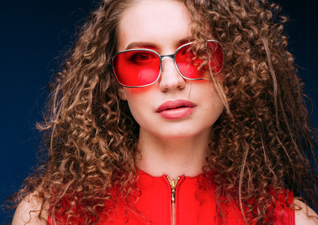 Beautiful attractive woman in red clothes and red fashion sun glasses over blue background and curly...