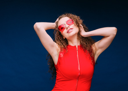 Beautiful attractive woman in red clothes and red fashion sun glasses over blue background and curly...