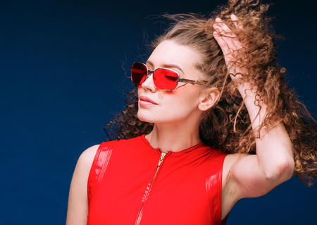 Beautiful attractive woman in red clothes and red fashion sun glasses over blue background and curly...