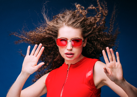 Beautiful attractive woman in red clothes and red fashion sun glasses over blue background and curly...