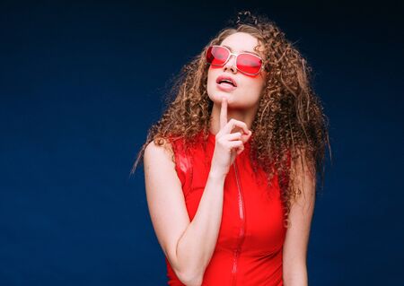 Beautiful attractive woman in red clothes and red fashion sun glasses over blue background and curly long beautiful hair. Studio shot.の写真素材
