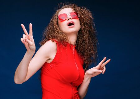 Beautiful attractive woman in red clothes and red fashion sun glasses over blue background and curly long beautiful hair. Studio shot.の写真素材