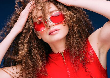 Beautiful attractive woman in red clothes and red fashion sun glasses over blue background and curly long beautiful hair. Studio shot.の写真素材