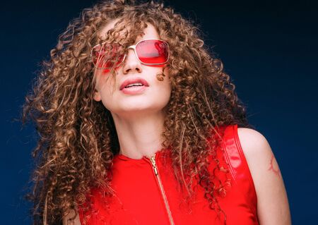 Beautiful attractive woman in red clothes and red fashion sun glasses over blue background and curly long beautiful hair. Studio shot.の写真素材
