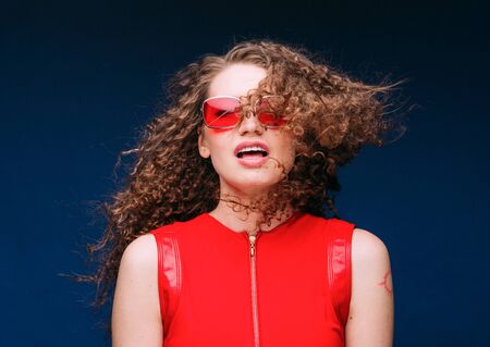 Beautiful attractive woman in red clothes and red fashion sun glasses over blue background and curly long beautiful hair. Studio shot.の写真素材