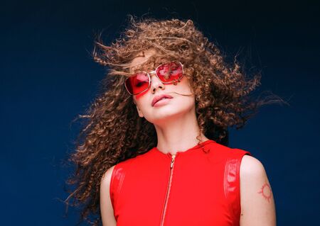Beautiful attractive woman in red clothes and red fashion sun glasses over blue background and curly long beautiful hair. Studio shot.の写真素材
