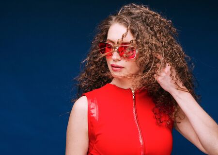 Beautiful attractive woman in red clothes and red fashion sun glasses over blue background and curly long beautiful hair. Studio shot.の写真素材