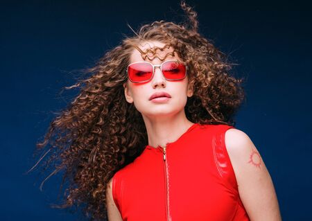 Beautiful attractive woman in red clothes and red fashion sun glasses over blue background and curly long beautiful hair. Studio shot.の写真素材
