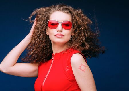Beautiful attractive woman in red clothes and red fashion sun glasses over blue background and curly long beautiful hair. Studio shot.の写真素材
