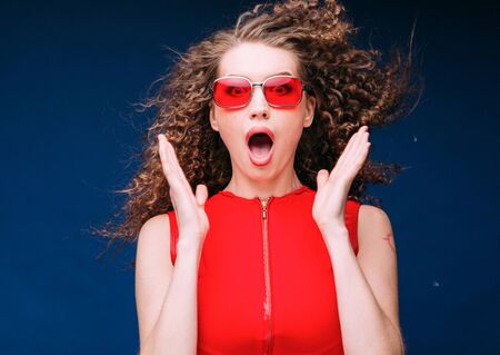 Beautiful attractive woman in red clothes and red fashion sun glasses over blue background and curly long beautiful hair. Studio shot.の写真素材