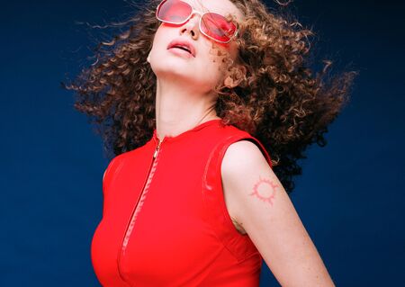 Beautiful attractive woman in red clothes and red fashion sun glasses over blue background and curly long beautiful hair. Studio shot.の写真素材