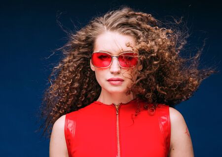 Beautiful attractive woman in red clothes and red fashion sun glasses over blue background and curly long beautiful hair. Studio shot.の写真素材
