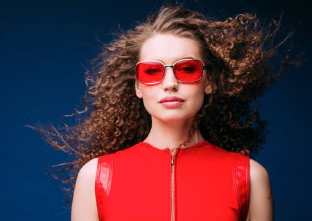 Beautiful attractive woman in red clothes and red fashion sun glasses over blue background and curly long beautiful hair. Studio shot.の写真素材