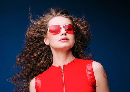 Beautiful attractive woman in red clothes and red fashion sun glasses over blue background and curly long beautiful hair. Studio shot.の写真素材