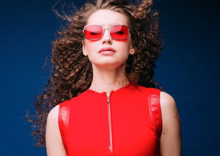 Beautiful attractive woman in red clothes and red fashion sun glasses over blue background and curly long beautiful hair. Studio shot.の写真素材