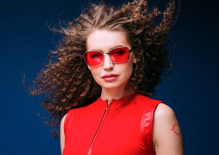 Beautiful attractive woman in red clothes and red fashion sun glasses over blue background and curly long beautiful hair. Studio shot.の写真素材