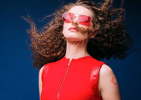 Beautiful attractive woman in red clothes and red fashion sun glasses over blue background and curly long beautiful hair. Studio shot.の写真素材