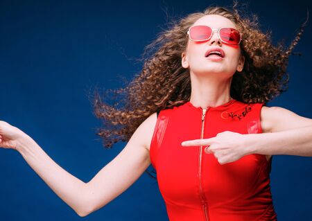 Beautiful attractive woman in red clothes and red fashion sun glasses over blue background and curly long beautiful hair. Studio shot.の写真素材