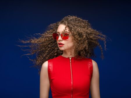 Beautiful attractive woman in red clothes and red fashion sun glasses over blue background and curly long beautiful hair. Studio shot.の写真素材