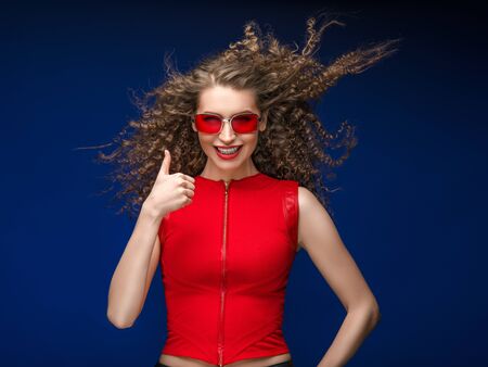 Beautiful attractive woman in red clothes and red fashion sun glasses over blue background and curly long beautiful hair. Studio shot.の写真素材