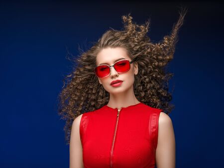 Beautiful attractive woman in red clothes and red fashion sun glasses over blue background and curly long beautiful hair. Studio shot.の写真素材