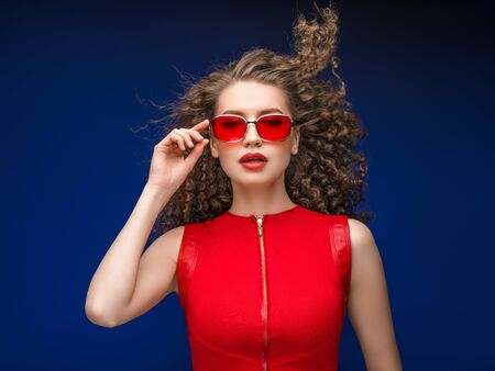 Beautiful attractive woman in red clothes and red fashion sun glasses over blue background and curly long beautiful hair. Studio shot.の写真素材