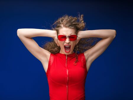 Beautiful attractive woman in red clothes and red fashion sun glasses over blue background and curly long beautiful hair. Studio shot.の写真素材
