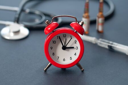 Red alarm clock in front of blurred stethoscope, syringe and medicine vials.の写真素材
