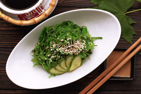 Chuka salad with cucumbers, sesame seeds and sauce, in a white plate, with a Japanese teapot, chopsticks and maple leaves, on a wooden backgroundの写真素材