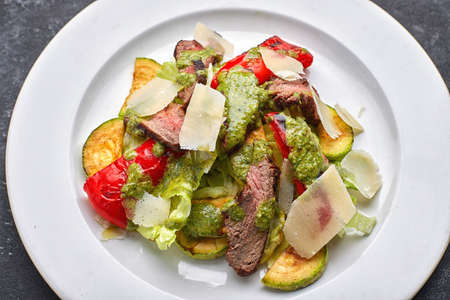 Warm salad with veal, baked vegetables, bell pepper, zucchini and parrmesan cheese, on a white plate, against a dark backgroundの写真素材