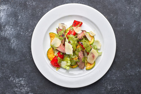 Warm salad with veal, baked vegetables, bell pepper, zucchini and parrmesan cheese, on a white plate, against a dark backgroundの写真素材