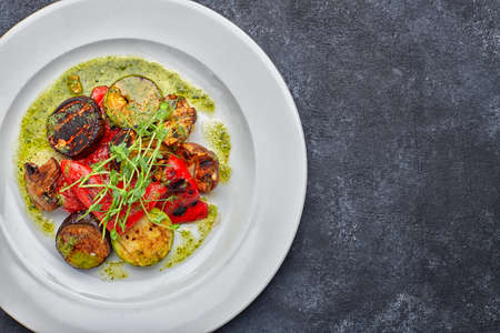 Grilled baked vegetables, eggplant, zucchini, bell pepper, champignon mushrooms, on a white plate against a dark background, with microgreenの写真素材